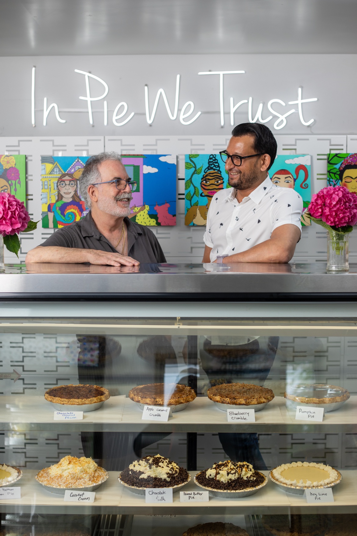 The owners of the Sweet and Savory Pie Company laughing together behind the pie case under the In Pie We Trust neon sign