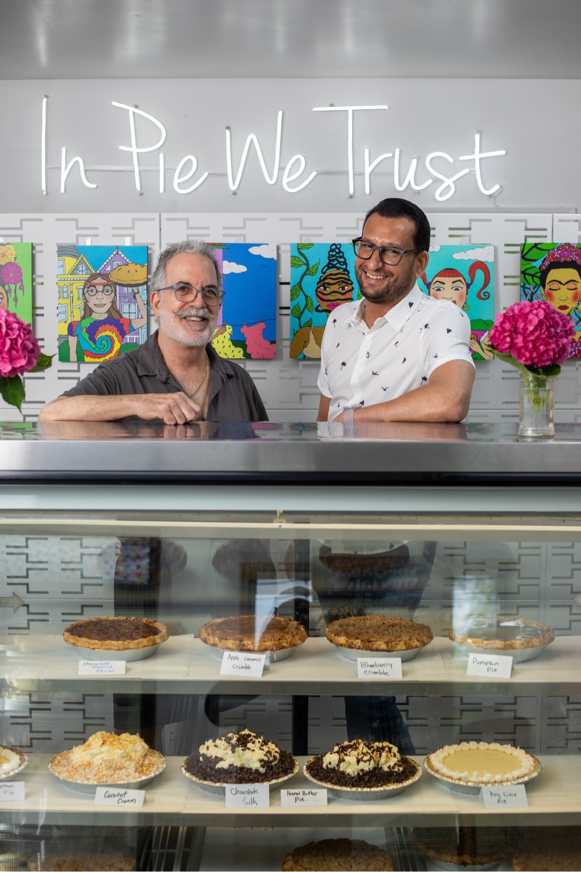 The bakery's owners standing behind the pie display case under the In Pie We Trust neon sign, with labeled fruit and cream pies in the case