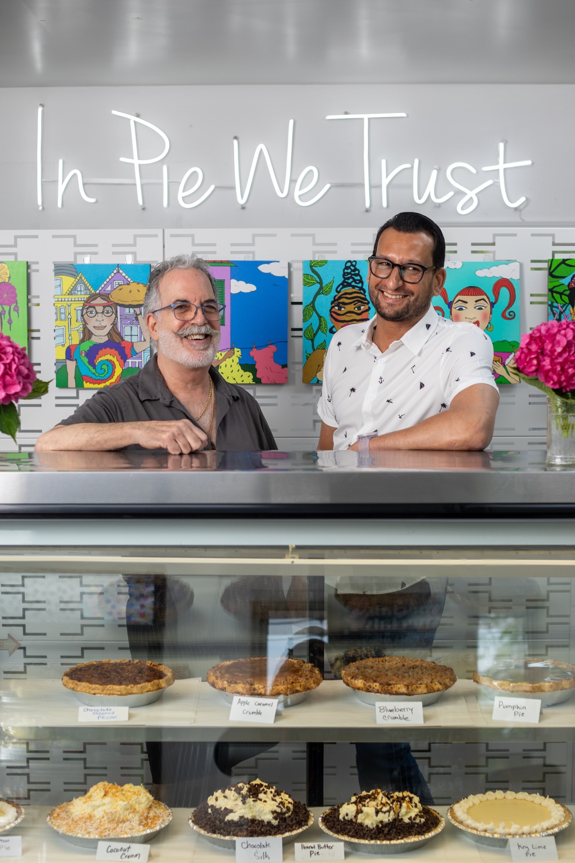 The owners smiling behind the pie case with labeled pies on the shelves below